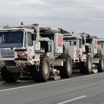 Mehrere weiße Vibro-Trucks stehen auf einer Straße am Rand eines Feldes. Die Spezialfahrzeuge werden für seismische Messungen zur Untersuchung des Untergrunds eingesetzt. Der Himmel ist bewölkt.