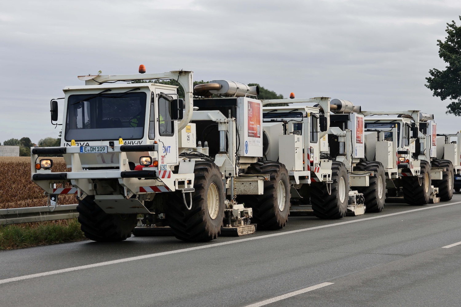Mehrere weiße Vibro-Trucks stehen auf einer Straße am Rand eines Feldes. Die Spezialfahrzeuge werden für seismische Messungen zur Untersuchung des Untergrunds eingesetzt. Der Himmel ist bewölkt.