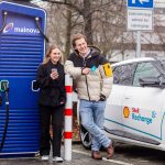 Zwei Menschen stehen an einer Ladesäule von Mainova neben einem Elektroauto mit Shell-Recharge-Logo. Die Frau hält ein Smartphone in der Hand, der Mann lehnt am Fahrzeug und zeigt eine Ladekarte. Die Szene zeigt das Laden eines Elektroautos im öffentlichen Raum.
