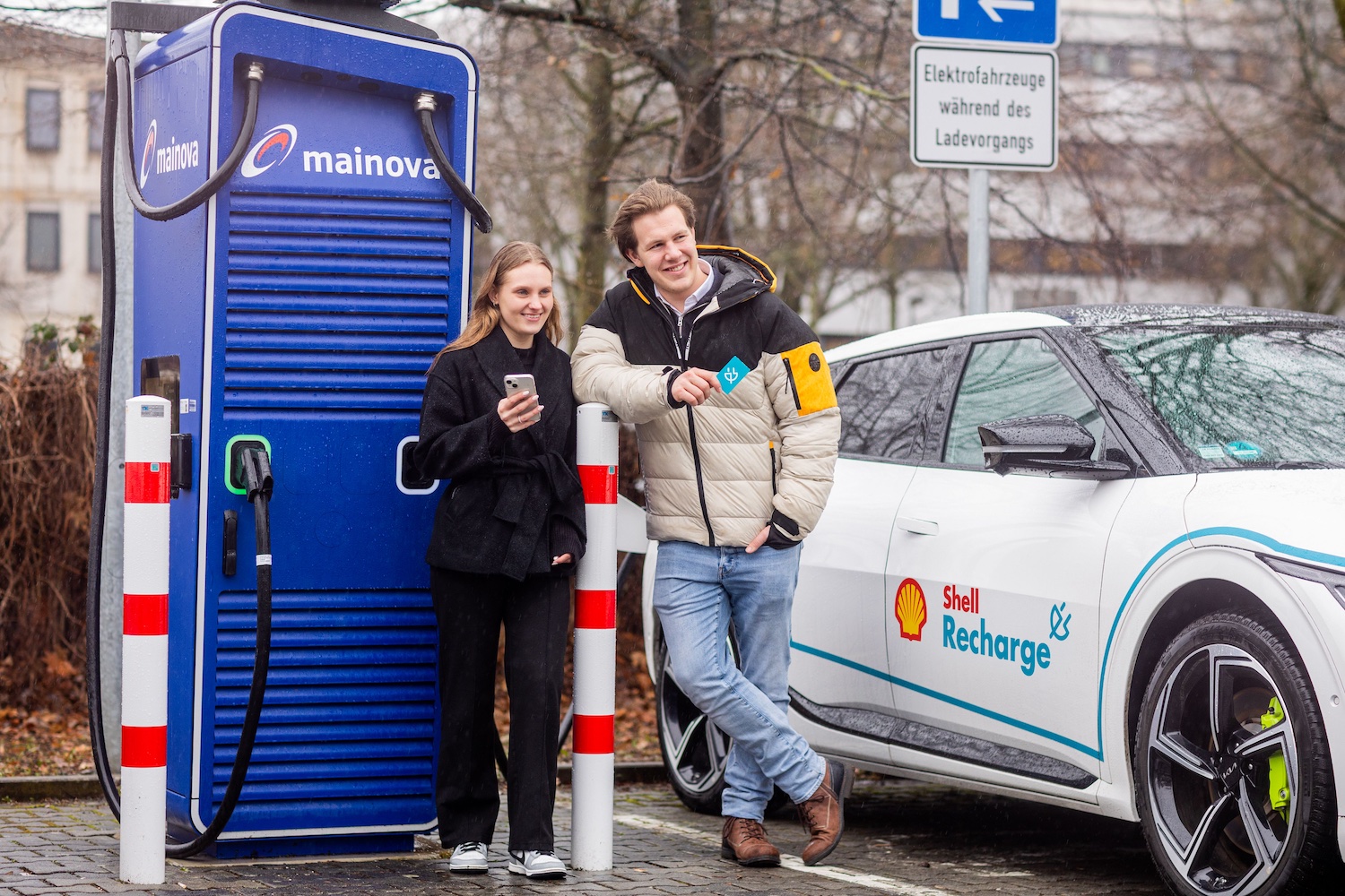Zwei Menschen stehen an einer Ladesäule von Mainova neben einem Elektroauto mit Shell-Recharge-Logo. Die Frau hält ein Smartphone in der Hand, der Mann lehnt am Fahrzeug und zeigt eine Ladekarte. Die Szene zeigt das Laden eines Elektroautos im öffentlichen Raum.