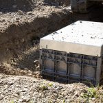 „Unterirdischer Einbau eines rechteckigen Kabelschachts in einer Baugrube. Der graue Kunststoffschacht mit Metallabdeckung wird in sandigem Erdreich installiert, während im Hintergrund ein Bagger zu sehen ist.“