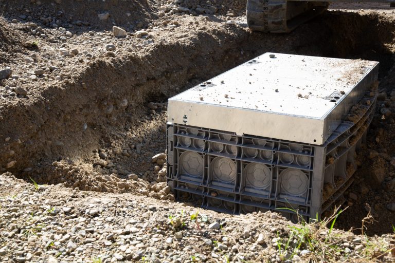 „Unterirdischer Einbau eines rechteckigen Kabelschachts in einer Baugrube. Der graue Kunststoffschacht mit Metallabdeckung wird in sandigem Erdreich installiert, während im Hintergrund ein Bagger zu sehen ist.“