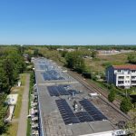 Luftaufnahme eines langen Gebäudes mit zahlreichen Solarmodulen auf dem Flachdach. Neben dem Gebäude verlaufen Bahngleise, umgeben von Bäumen und Wohnhäusern. Im Hintergrund sind weitere Gebäude und viel Grünfläche unter klarem blauem Himmel zu sehen.