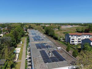 Luftaufnahme eines langen Gebäudes mit zahlreichen Solarmodulen auf dem Flachdach. Neben dem Gebäude verlaufen Bahngleise, umgeben von Bäumen und Wohnhäusern. Im Hintergrund sind weitere Gebäude und viel Grünfläche unter klarem blauem Himmel zu sehen.
