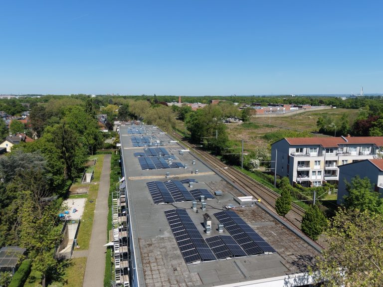 Luftaufnahme eines langen Gebäudes mit zahlreichen Solarmodulen auf dem Flachdach. Neben dem Gebäude verlaufen Bahngleise, umgeben von Bäumen und Wohnhäusern. Im Hintergrund sind weitere Gebäude und viel Grünfläche unter klarem blauem Himmel zu sehen.
