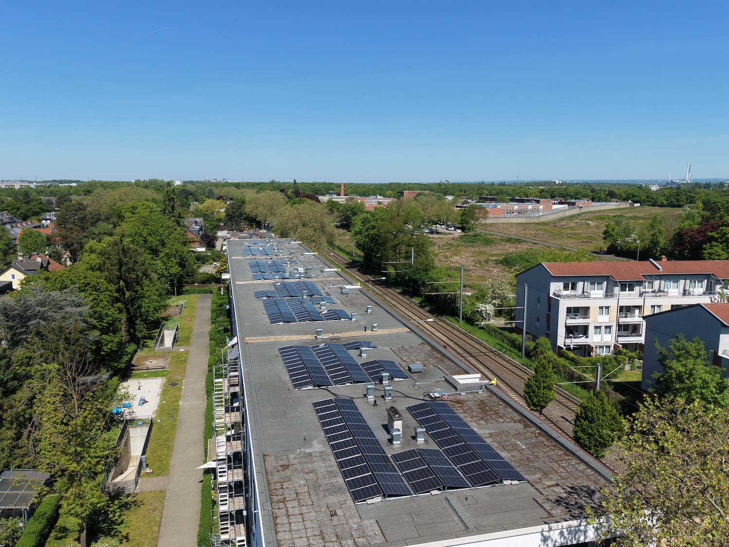 Luftaufnahme eines langen Gebäudes mit zahlreichen Solarmodulen auf dem Flachdach. Neben dem Gebäude verlaufen Bahngleise, umgeben von Bäumen und Wohnhäusern. Im Hintergrund sind weitere Gebäude und viel Grünfläche unter klarem blauem Himmel zu sehen.