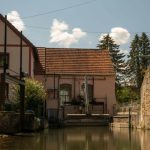 Historisches Gebäude mit rotem Ziegeldach an einem ruhigen Wasserkanal, davor technische Anlagen und Schleusenelemente; im Hintergrund stehen hohe Nadelbäume unter blauem Himmel mit einzelnen Wolken.