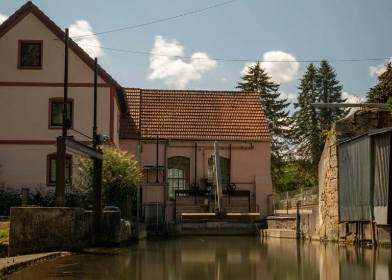 Historisches Gebäude mit rotem Ziegeldach an einem ruhigen Wasserkanal, davor technische Anlagen und Schleusenelemente; im Hintergrund stehen hohe Nadelbäume unter blauem Himmel mit einzelnen Wolken.