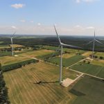 Luftaufnahme eines Windparks in einer ländlichen Landschaft. Drei große Windenergieanlagen stehen zwischen Feldern, Wiesen und Baumreihen. Die Rotorblätter werfen lange Schatten auf die Äcker. Im Hintergrund sind Wälder und ein weiter Horizont unter leicht bewölktem Himmel zu sehen.