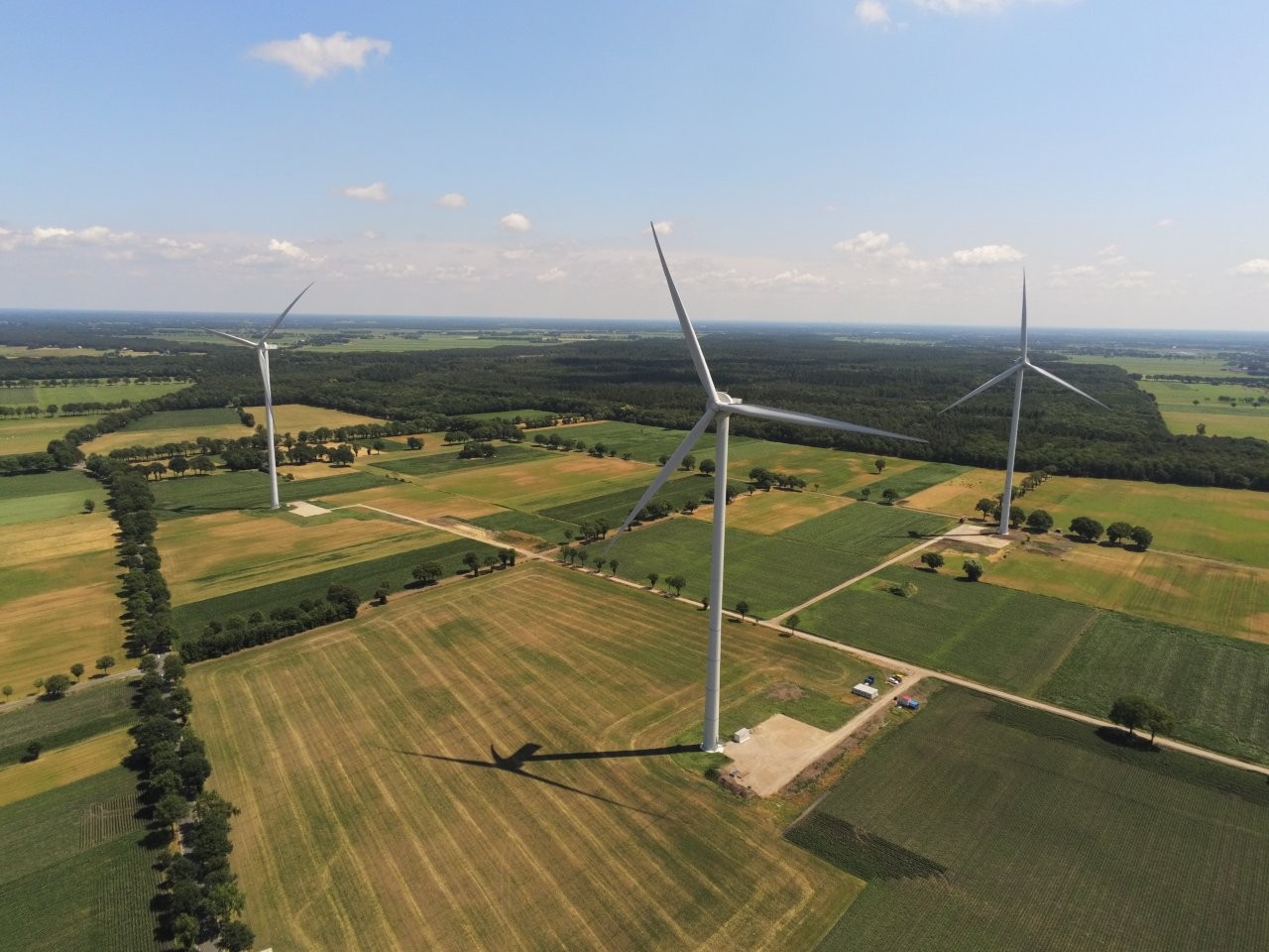 Luftaufnahme eines Windparks in einer ländlichen Landschaft. Drei große Windenergieanlagen stehen zwischen Feldern, Wiesen und Baumreihen. Die Rotorblätter werfen lange Schatten auf die Äcker. Im Hintergrund sind Wälder und ein weiter Horizont unter leicht bewölktem Himmel zu sehen.