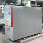 Blockheizkraftwerk in einem Technikraum: graues, rechteckiges Metallgehäuse mit rotem Seitenstreifen, Bedienfeld und Not-Aus-Schalter an der Front, oben angeschlossene Kabel und Leitungen, installiert auf einem Betonsockel.