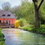 Kleines Wasserkraftwerk an einem schmalen Fluss: Ein Gebäude mit roter Fassade und Satteldach steht direkt am Ufer, daneben ist eine Anlage zur Wasserführung erkennbar. Der Fluss fließt ruhig durch eine grüne, bewaldete Landschaft, im Vordergrund schwimmt eine Ente.