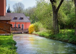 Kleines Wasserkraftwerk an einem schmalen Fluss: Ein Gebäude mit roter Fassade und Satteldach steht direkt am Ufer, daneben ist eine Anlage zur Wasserführung erkennbar. Der Fluss fließt ruhig durch eine grüne, bewaldete Landschaft, im Vordergrund schwimmt eine Ente.