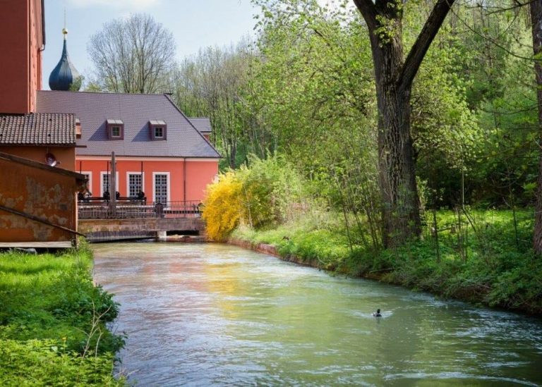 Kleines Wasserkraftwerk an einem schmalen Fluss: Ein Gebäude mit roter Fassade und Satteldach steht direkt am Ufer, daneben ist eine Anlage zur Wasserführung erkennbar. Der Fluss fließt ruhig durch eine grüne, bewaldete Landschaft, im Vordergrund schwimmt eine Ente.