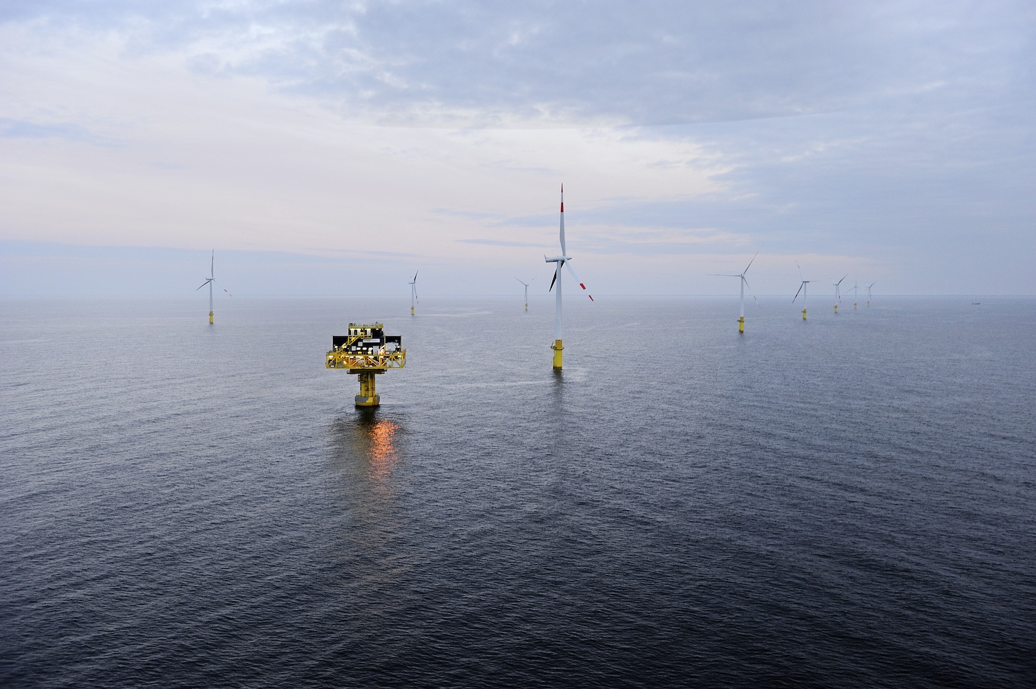 Offshore-Windpark Baltic 1 in der Ostsee: Mehrere Windkraftanlagen stehen in gleichmäßigen Abständen im offenen Meer. Im Vordergrund ist eine Plattform für Technik oder Wartung zu sehen, dahinter drehen sich die Rotoren der Windräder unter einem bewölkten Himmel.