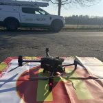 Eine Drohne steht auf einem rot-weiß markierten Landeplatz am Straßenrand vor einem Servicefahrzeug von WEMAG Netz. Das Fahrzeug ist mit einer Leiter ausgestattet und parkt neben einer ländlichen Straße mit kahlen Bäumen und freiem Feld im Hintergrund. Die Szene wirkt sonnig und zeigt den Einsatz moderner Drohnentechnik zur Kontrolle von Stromnetzen.