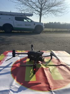 Eine Drohne steht auf einem rot-weiß markierten Landeplatz am Straßenrand vor einem Servicefahrzeug von WEMAG Netz. Das Fahrzeug ist mit einer Leiter ausgestattet und parkt neben einer ländlichen Straße mit kahlen Bäumen und freiem Feld im Hintergrund. Die Szene wirkt sonnig und zeigt den Einsatz moderner Drohnentechnik zur Kontrolle von Stromnetzen.