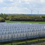 Große Solarthermieanlage mit Reihen von Parabolrinnenkollektoren auf einer grünen Fläche. Im Hintergrund stehen mehrere Windräder vor einer bewaldeten Landschaft unter bewölktem Himmel.