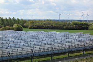 Große Solarthermieanlage mit Reihen von Parabolrinnenkollektoren auf einer grünen Fläche. Im Hintergrund stehen mehrere Windräder vor einer bewaldeten Landschaft unter bewölktem Himmel.