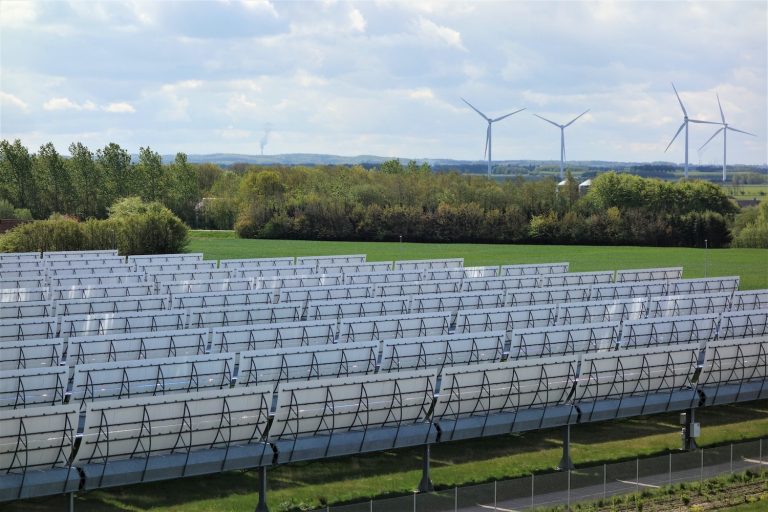 Große Solarthermieanlage mit Reihen von Parabolrinnenkollektoren auf einer grünen Fläche. Im Hintergrund stehen mehrere Windräder vor einer bewaldeten Landschaft unter bewölktem Himmel.