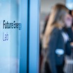 nscharfe Aufnahme eines modernen Büro- oder Veranstaltungsraums. Im Vordergrund ist auf einer Glasfläche der Schriftzug „Future Energy Lab“ zu sehen. Im Hintergrund stehen mehrere verschwommene Personen an einem Stehtisch im Gespräch.