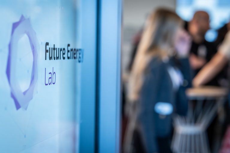 nscharfe Aufnahme eines modernen Büro- oder Veranstaltungsraums. Im Vordergrund ist auf einer Glasfläche der Schriftzug „Future Energy Lab“ zu sehen. Im Hintergrund stehen mehrere verschwommene Personen an einem Stehtisch im Gespräch.