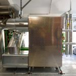 Große silberne Rohrleitungen und technische Anlagen in einem Heizungs- oder Energietechnikraum. Im Mittelpunkt steht ein rechteckiges Metallgehäuse mit strukturierter Oberfläche, umgeben von isolierten Rohren, Ventilen und Messinstrumenten.