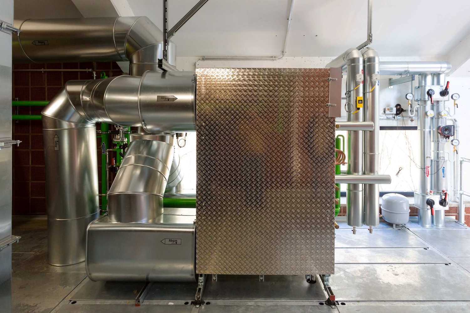 Große silberne Rohrleitungen und technische Anlagen in einem Heizungs- oder Energietechnikraum. Im Mittelpunkt steht ein rechteckiges Metallgehäuse mit strukturierter Oberfläche, umgeben von isolierten Rohren, Ventilen und Messinstrumenten.