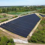 Luftaufnahme einer großen Freiflächen-Photovoltaikanlage mit zahlreichen dunkelblauen Solarmodulen, eingebettet in eine grüne Landschaft nahe einer Bahnstrecke und landwirtschaftlichen Flächen. Im Hintergrund ist eine Stadt mit Wohn- und Industriegebäuden zu sehen.