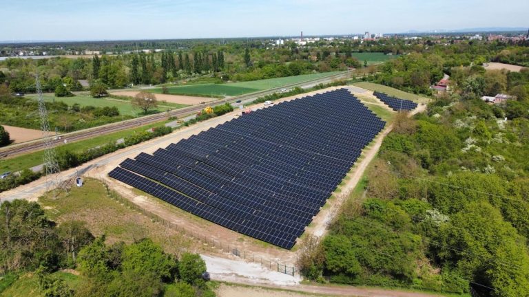Luftaufnahme einer großen Freiflächen-Photovoltaikanlage mit zahlreichen dunkelblauen Solarmodulen, eingebettet in eine grüne Landschaft nahe einer Bahnstrecke und landwirtschaftlichen Flächen. Im Hintergrund ist eine Stadt mit Wohn- und Industriegebäuden zu sehen.