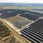 „Luftaufnahme eines großflächigen Solarparks mit langen Reihen dunkelblauer Solarmodule auf einer offenen Landschaftsfläche. Die Solarfelder erstrecken sich über mehrere Bereiche und sind von Feldwegen, Wiesen und kleinen Waldstücken umgeben. Im Hintergrund sind Felder, Wälder und mehrere Windkraftanlagen am Horizont zu sehen.“