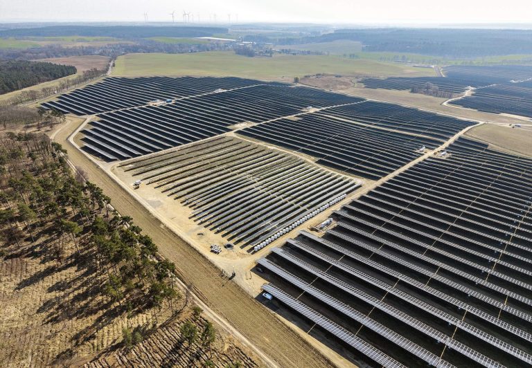 „Luftaufnahme eines großflächigen Solarparks mit langen Reihen dunkelblauer Solarmodule auf einer offenen Landschaftsfläche. Die Solarfelder erstrecken sich über mehrere Bereiche und sind von Feldwegen, Wiesen und kleinen Waldstücken umgeben. Im Hintergrund sind Felder, Wälder und mehrere Windkraftanlagen am Horizont zu sehen.“