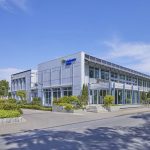 Modernes Gebäude der Stadtwerke Hilden mit großer Glasfassade und silberner Metallverkleidung an einer ruhigen Straße bei sonnigem Wetter. Vor dem Eingangsbereich befinden sich Grünanlagen, kleine Bäume und ein gepflasterter Gehweg.