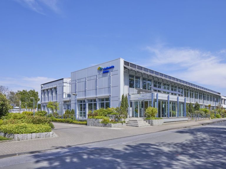 Modernes Gebäude der Stadtwerke Hilden mit großer Glasfassade und silberner Metallverkleidung an einer ruhigen Straße bei sonnigem Wetter. Vor dem Eingangsbereich befinden sich Grünanlagen, kleine Bäume und ein gepflasterter Gehweg.