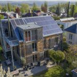 Mehrstöckiges Wohnhaus mit Holzfassade und großer Photovoltaikanlage auf dem Dach in einem grünen Wohngebiet. Das Gebäude verfügt über Balkone, große Fenster und eine Terrasse mit Sitzmöbeln. Im Hintergrund sind weitere Häuser, Bäume und eine hügelige Landschaft zu sehen.