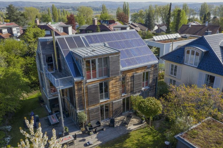 Mehrstöckiges Wohnhaus mit Holzfassade und großer Photovoltaikanlage auf dem Dach in einem grünen Wohngebiet. Das Gebäude verfügt über Balkone, große Fenster und eine Terrasse mit Sitzmöbeln. Im Hintergrund sind weitere Häuser, Bäume und eine hügelige Landschaft zu sehen.