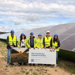 Fünf Personen mit Schutzhelmen und Warnwesten stehen vor einer großen Freiflächen-Solaranlage. Sie halten ein Schild mit der Aufschrift „Hier entsteht der Solarpark Auhofen“. Im Hintergrund sind lange Reihen von Solarmodulen unter einem bewölkten Himmel zu sehen.
