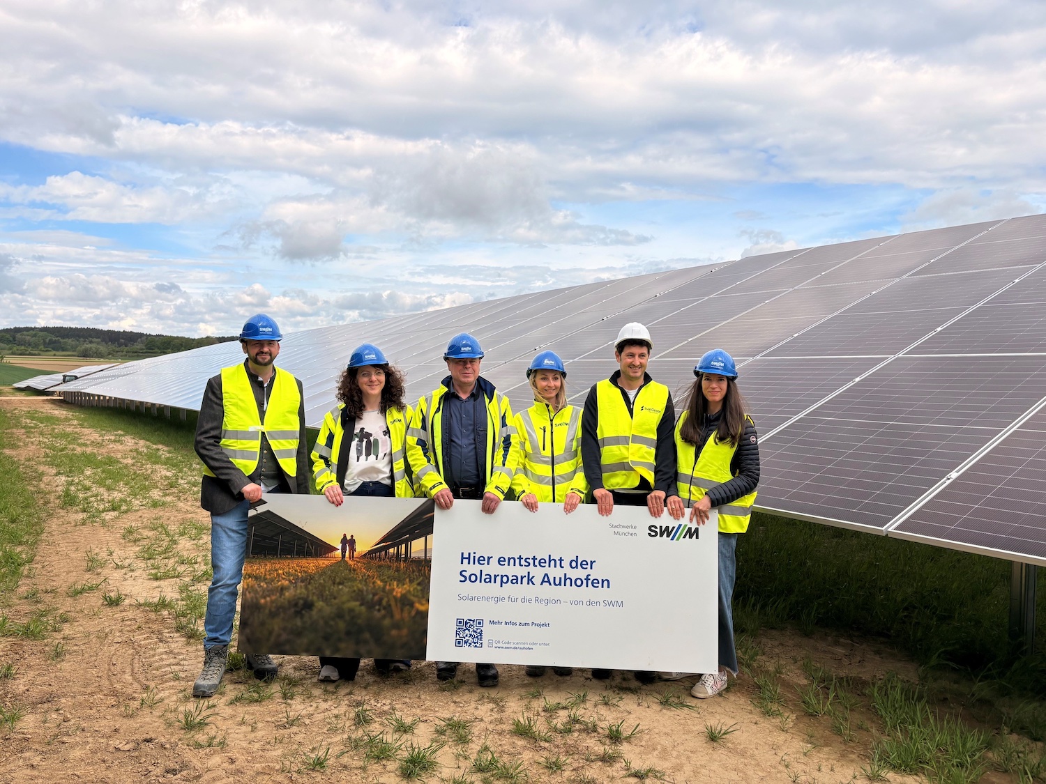 Fünf Personen mit Schutzhelmen und Warnwesten stehen vor einer großen Freiflächen-Solaranlage. Sie halten ein Schild mit der Aufschrift „Hier entsteht der Solarpark Auhofen“. Im Hintergrund sind lange Reihen von Solarmodulen unter einem bewölkten Himmel zu sehen.
