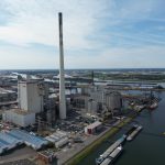 Luftaufnahme eines großen Kraftwerks im Industriehafen von Bremen: Mehrere Industriegebäude mit einem sehr hohen, zentralen Schornstein dominieren das Gelände. Das Kraftwerk liegt direkt am Wasser, an dessen Ufer Schiffe und Lastkähne angelegt sind. Im Hintergrund erstrecken sich weitere Hafenanlagen, Lagerflächen und vereinzelte Windkraftanlagen unter einem leicht bewölkten Himmel.