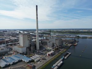 Luftaufnahme eines großen Kraftwerks im Industriehafen von Bremen: Mehrere Industriegebäude mit einem sehr hohen, zentralen Schornstein dominieren das Gelände. Das Kraftwerk liegt direkt am Wasser, an dessen Ufer Schiffe und Lastkähne angelegt sind. Im Hintergrund erstrecken sich weitere Hafenanlagen, Lagerflächen und vereinzelte Windkraftanlagen unter einem leicht bewölkten Himmel.