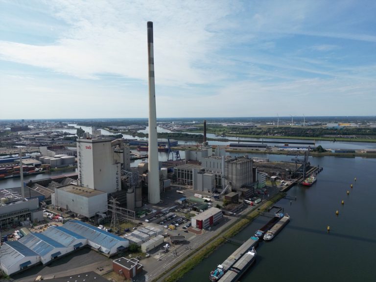 Luftaufnahme eines großen Kraftwerks im Industriehafen von Bremen: Mehrere Industriegebäude mit einem sehr hohen, zentralen Schornstein dominieren das Gelände. Das Kraftwerk liegt direkt am Wasser, an dessen Ufer Schiffe und Lastkähne angelegt sind. Im Hintergrund erstrecken sich weitere Hafenanlagen, Lagerflächen und vereinzelte Windkraftanlagen unter einem leicht bewölkten Himmel.