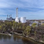 Blick auf den Energiestandort Süd der Stadtwerke München mit dem neuen Wärmespeicher. Der große, zylindrische Speicher steht neben mehreren Schornsteinen des Heizkraftwerks am Ufer der Isar. Im Vordergrund sind Bäume und das Flussufer zu sehen, im Hintergrund Teile des Münchner Stadtgebiets.