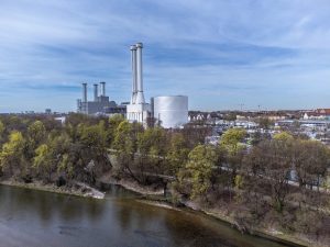 Blick auf den Energiestandort Süd der Stadtwerke München mit dem neuen Wärmespeicher. Der große, zylindrische Speicher steht neben mehreren Schornsteinen des Heizkraftwerks am Ufer der Isar. Im Vordergrund sind Bäume und das Flussufer zu sehen, im Hintergrund Teile des Münchner Stadtgebiets.