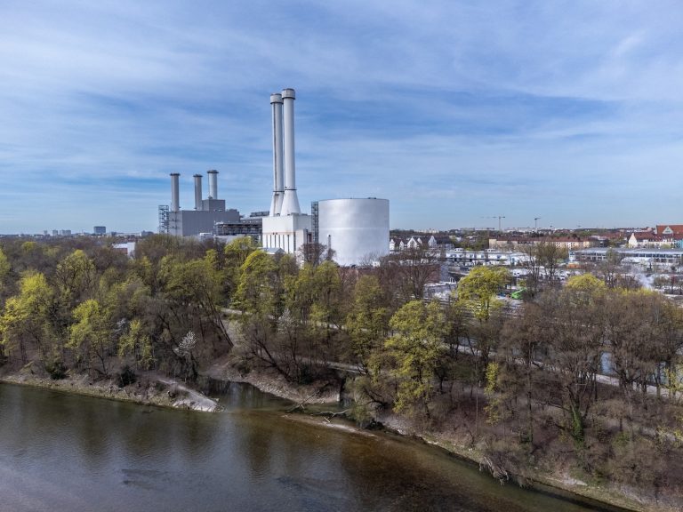 Blick auf den Energiestandort Süd der Stadtwerke München mit dem neuen Wärmespeicher. Der große, zylindrische Speicher steht neben mehreren Schornsteinen des Heizkraftwerks am Ufer der Isar. Im Vordergrund sind Bäume und das Flussufer zu sehen, im Hintergrund Teile des Münchner Stadtgebiets.