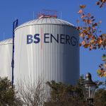 Zwei große, zylindrische Wärmespeicher mit dem Schriftzug „BS|ENERGY“ stehen hinter Bäumen unter blauem Himmel. Im Vordergrund sind herbstlich gefärbte Blätter und eine Straßenlaterne zu sehen.