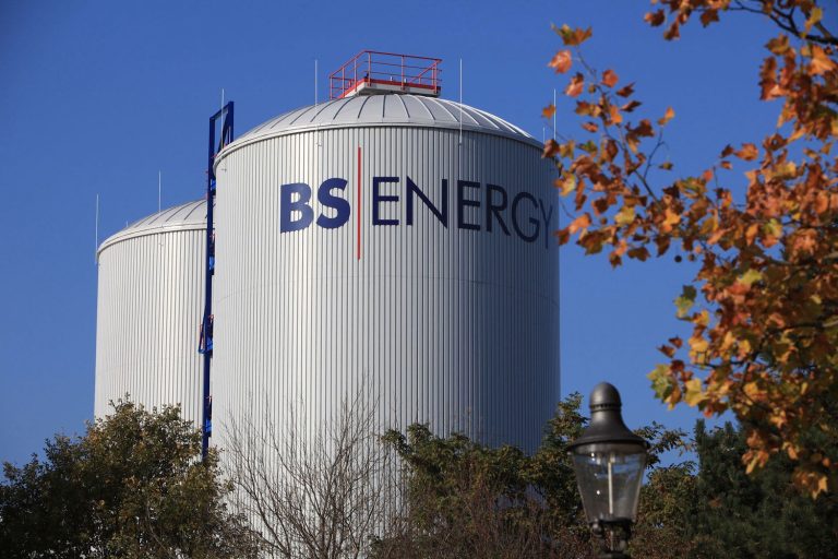 Zwei große, zylindrische Wärmespeicher mit dem Schriftzug „BS|ENERGY“ stehen hinter Bäumen unter blauem Himmel. Im Vordergrund sind herbstlich gefärbte Blätter und eine Straßenlaterne zu sehen.