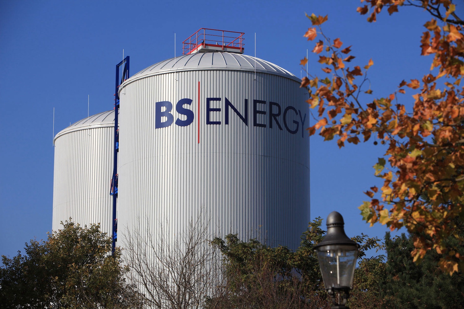 Zwei große, zylindrische Wärmespeicher mit dem Schriftzug „BS|ENERGY“ stehen hinter Bäumen unter blauem Himmel. Im Vordergrund sind herbstlich gefärbte Blätter und eine Straßenlaterne zu sehen.