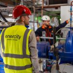 Ein Mitarbeiter von enercity trägt einen roten Schutzhelm und eine gelbe Warnweste und steht in einer technischen Anlage vor einer großen blauen Pumpeneinheit. Im Hintergrund bedient ein weiterer Mitarbeiter Armaturen und Rohrleitungen. Das Bild zeigt Arbeiten an einer Anlage zur Wärme- oder Energieversorgung.