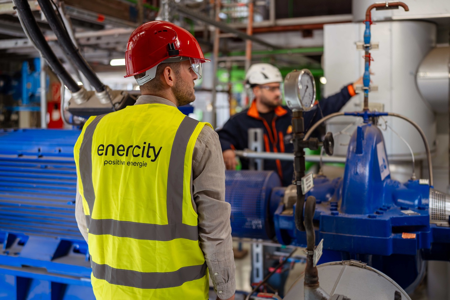 Ein Mitarbeiter von enercity trägt einen roten Schutzhelm und eine gelbe Warnweste und steht in einer technischen Anlage vor einer großen blauen Pumpeneinheit. Im Hintergrund bedient ein weiterer Mitarbeiter Armaturen und Rohrleitungen. Das Bild zeigt Arbeiten an einer Anlage zur Wärme- oder Energieversorgung.