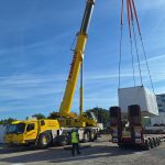 Ein Autokran hebt einen großen, weiß verpackten Batteriespeicher von einem Tieflader auf einer Baustelle im Energiepark Philippsburg. Im Vordergrund überwacht eine Person in Warnweste den Hebevorgang. Im Hintergrund sind ein Industriegebäude und weitere Baustellenfahrzeuge unter blauem Himmel zu sehen.